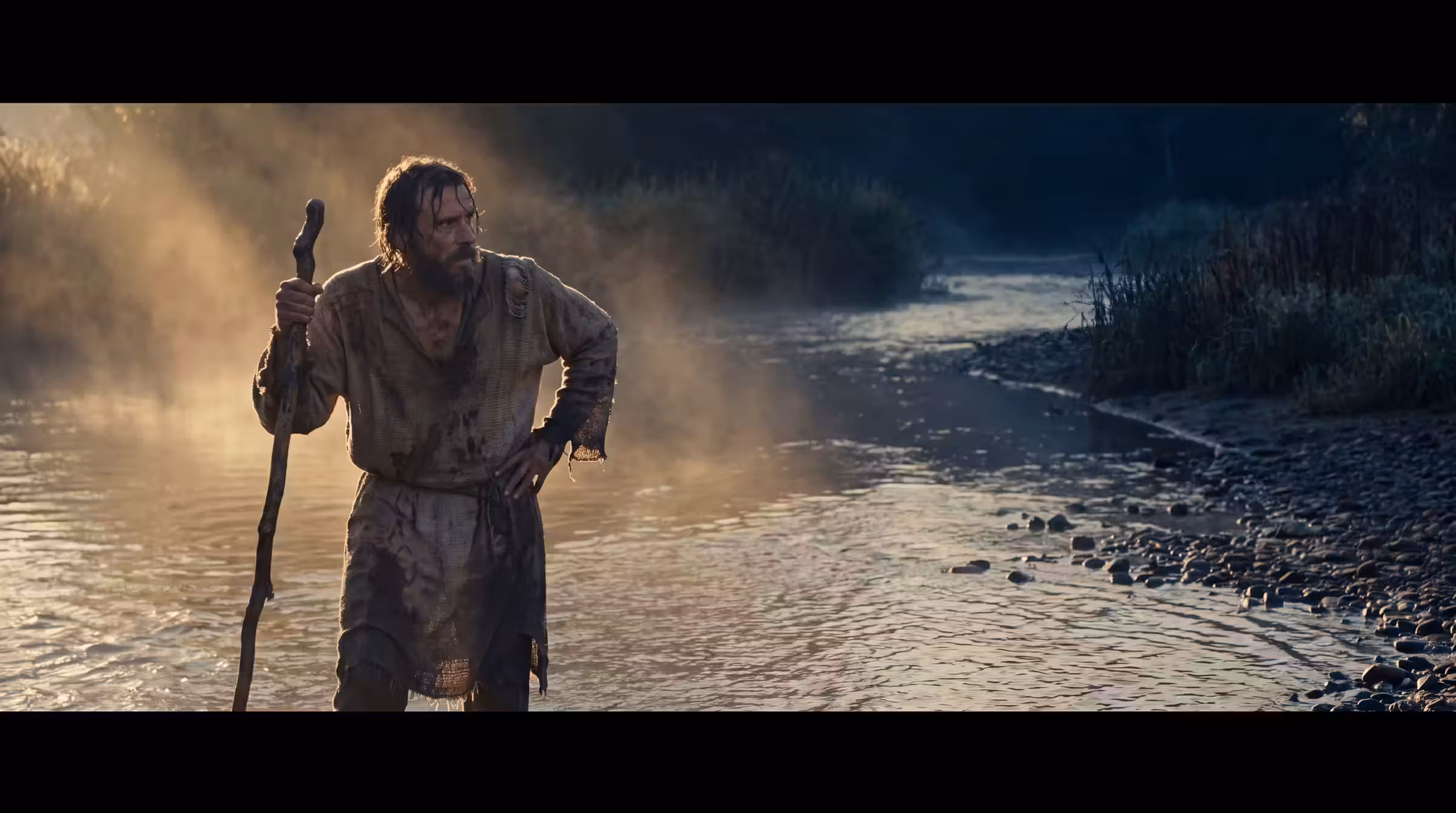 A limping man in a torn tunic leaning on a staff at a misty river ford at sunrise, looking back at the empty bank behind him