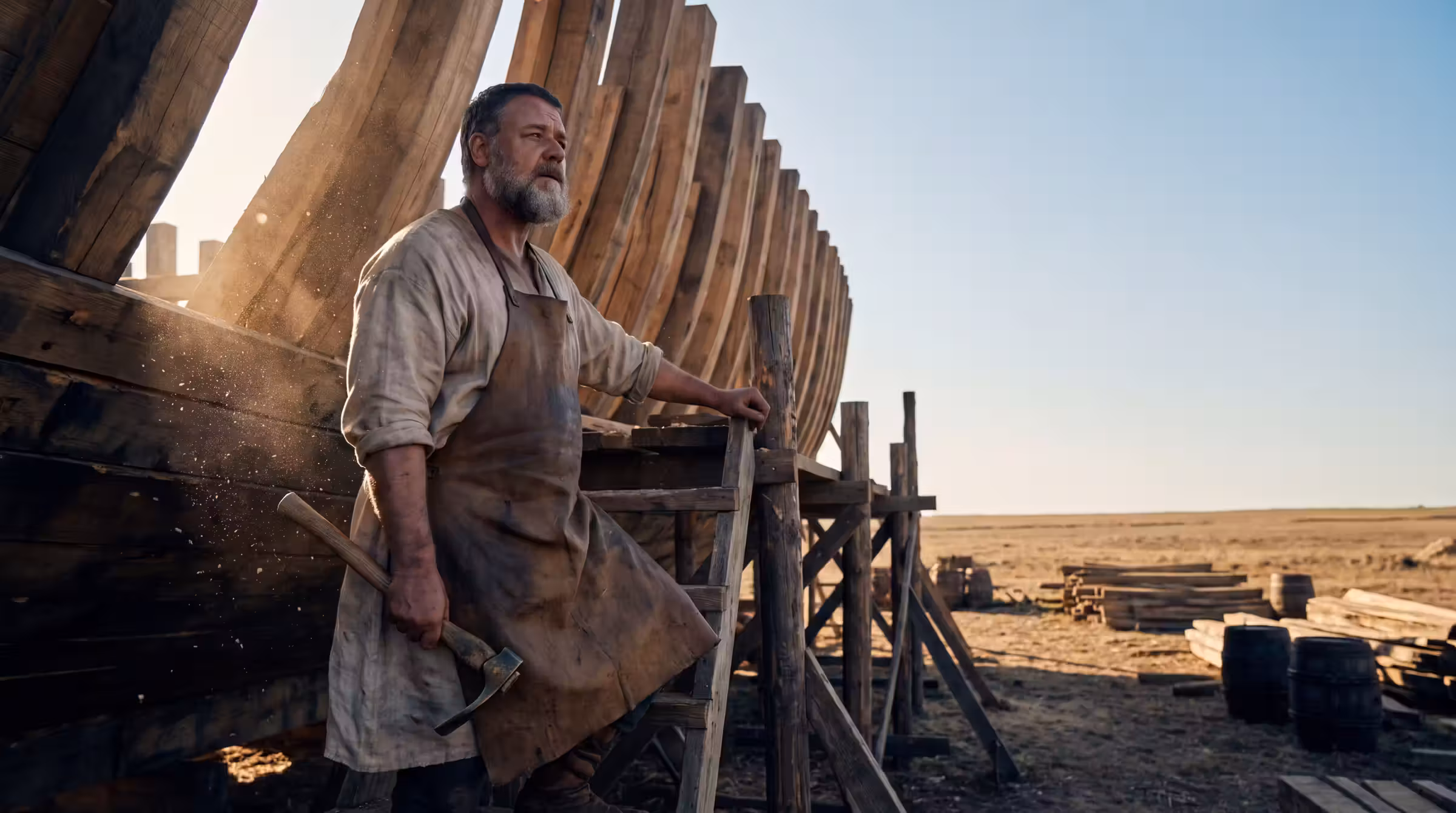 A bearded man with a bronze adze on scaffolding beside the ribbed timber hull of an enormous unfinished ship on a dry plain