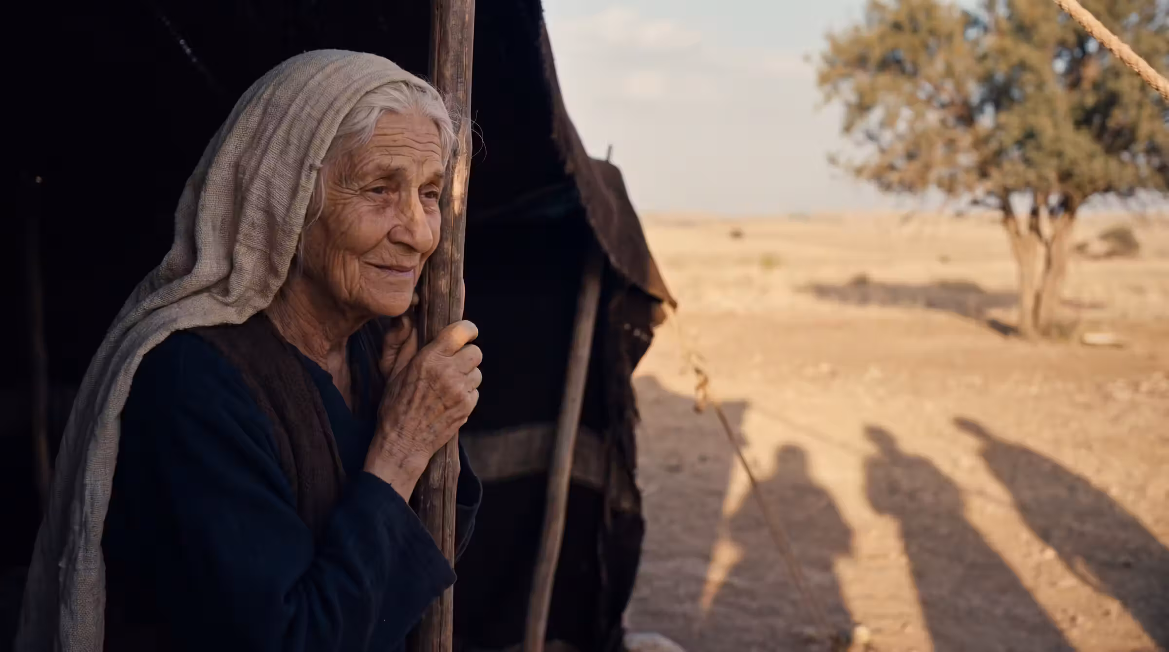 An elderly woman listening from inside the shadowed opening of a tent, a half-smile beginning, three long shadows on the sunlit ground outside