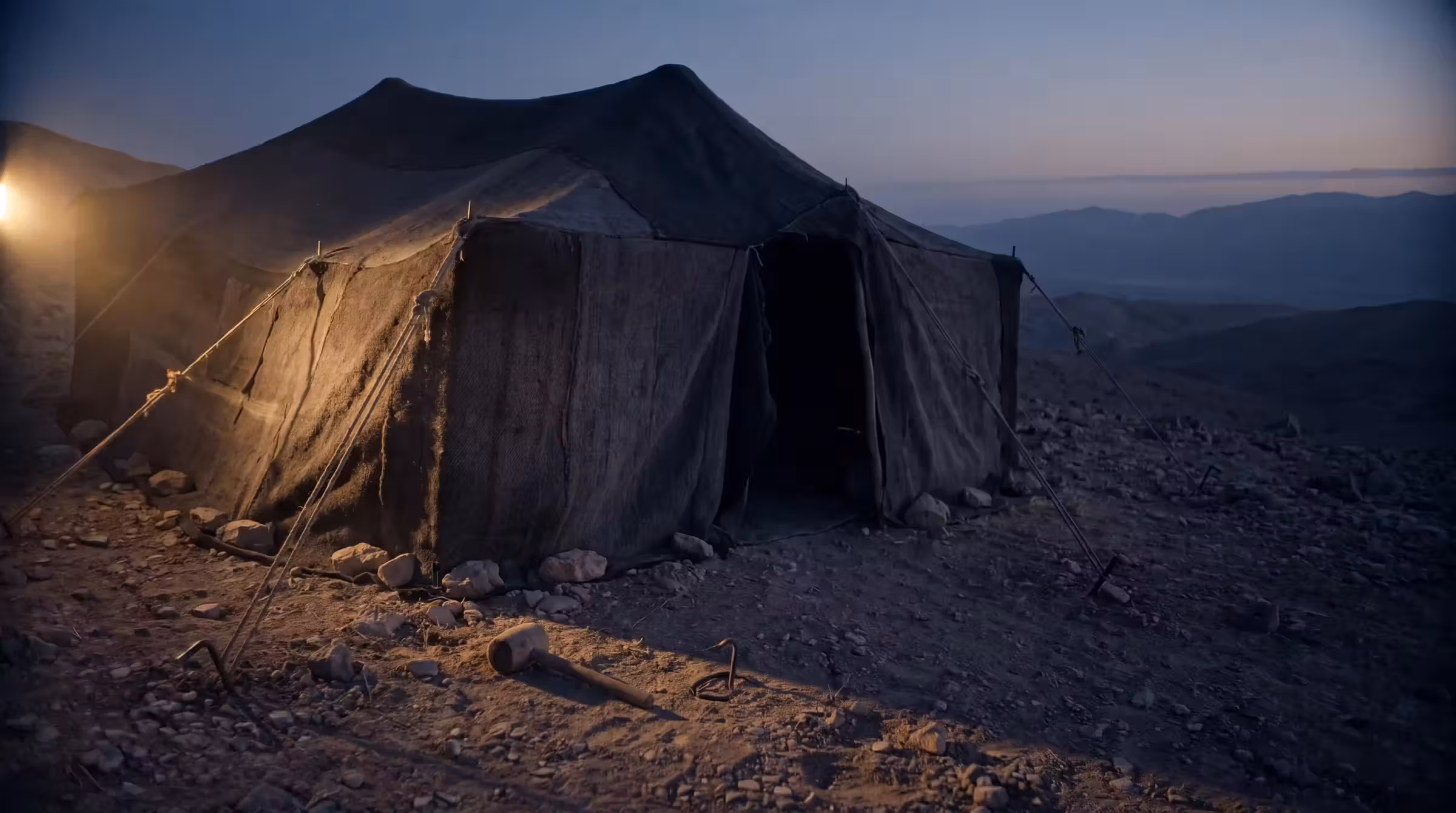 A dark goat-hair bedouin tent on a stony hillside at dusk, a wooden mallet and iron tent peg lying in the dust outside