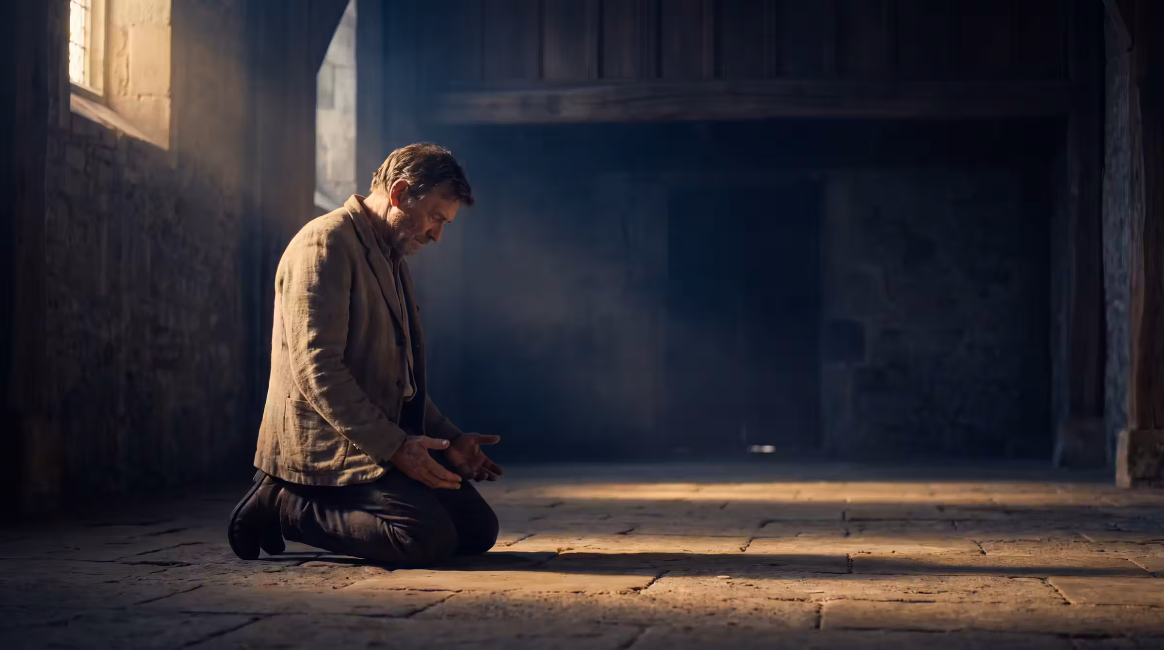 A kneeling man with upturned open hands in an empty stone chapel, lit by one shaft of morning light from a high window