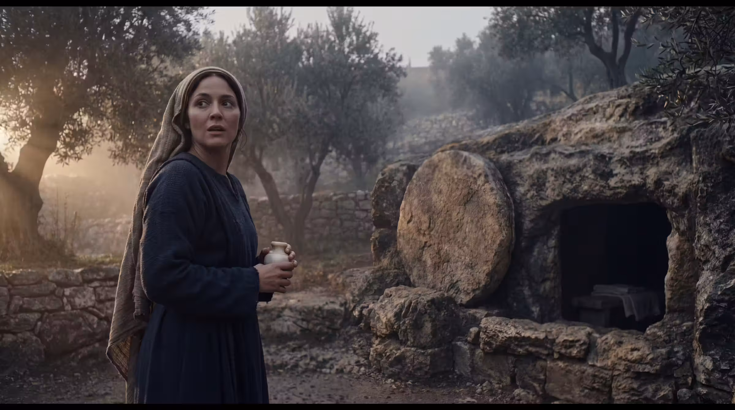 A woman holding an alabaster jar turning in a garden at sunrise, an open rock-cut tomb with its stone rolled clear behind her