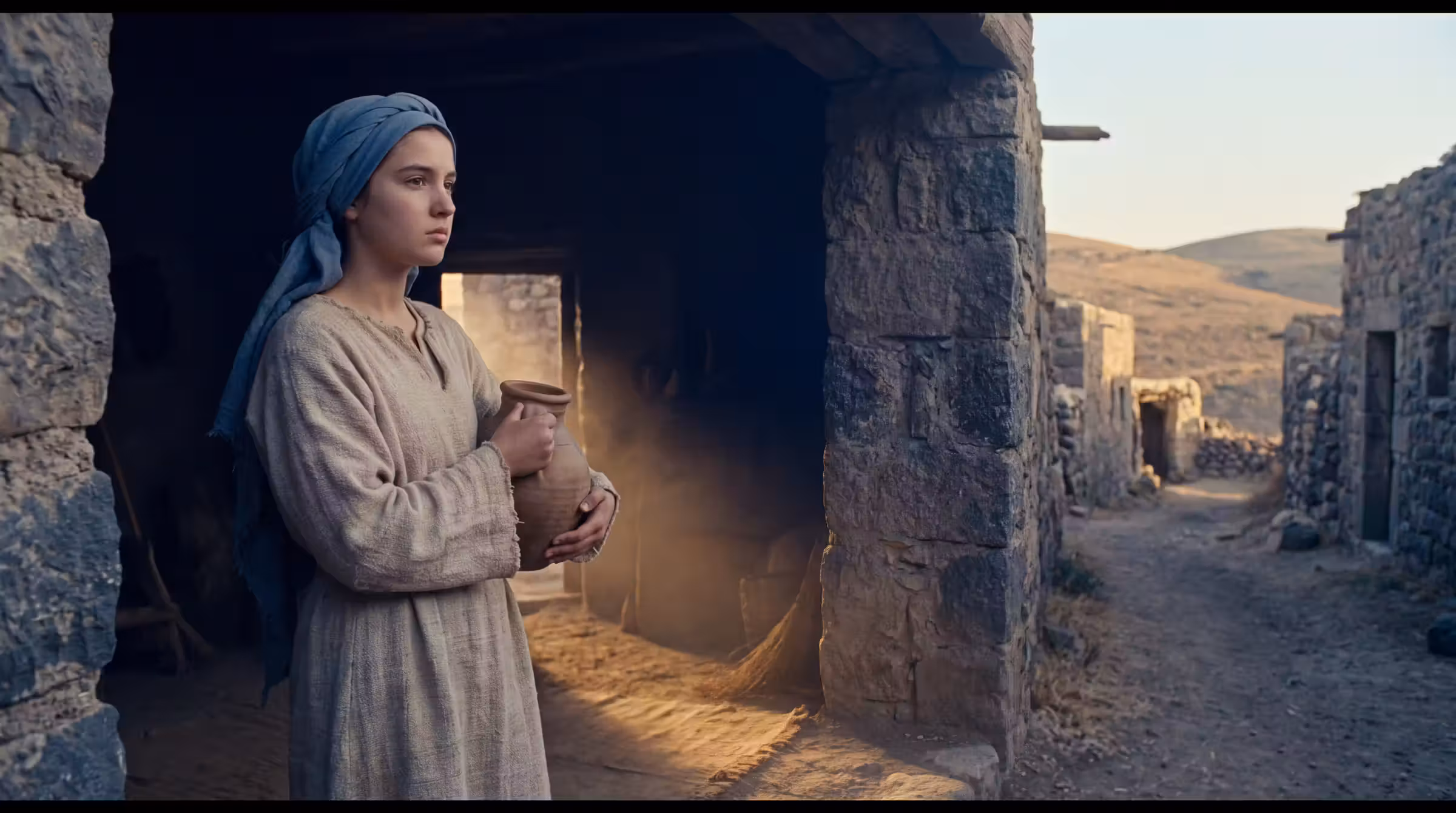 A teenage girl in a plain robe and blue head covering standing in the doorway of a small stone house holding a clay water jar