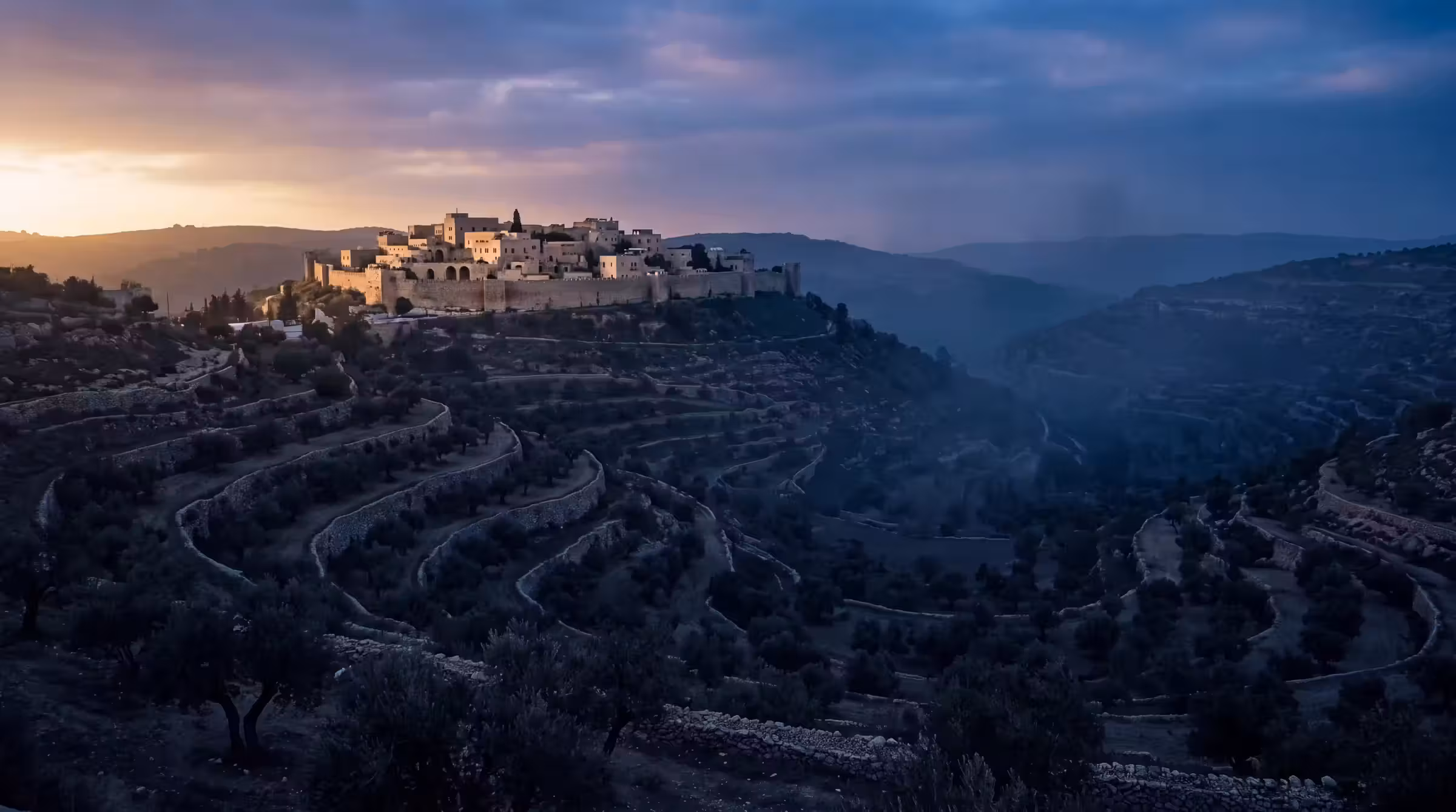 A Levantine valley at sundown with ancient dry-stone olive terraces and a walled hilltop town catching the last light